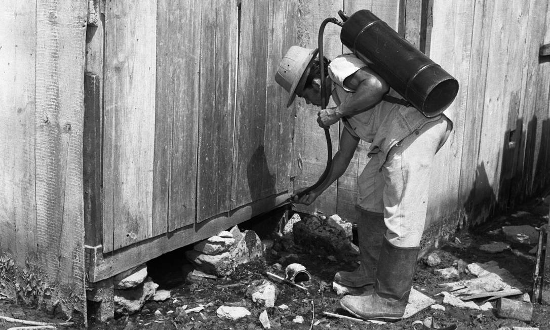 Guarda sanitário com pulverizador costal aplica inseticida na base de uma casa de madeira, em foto antiga em preto e branco
