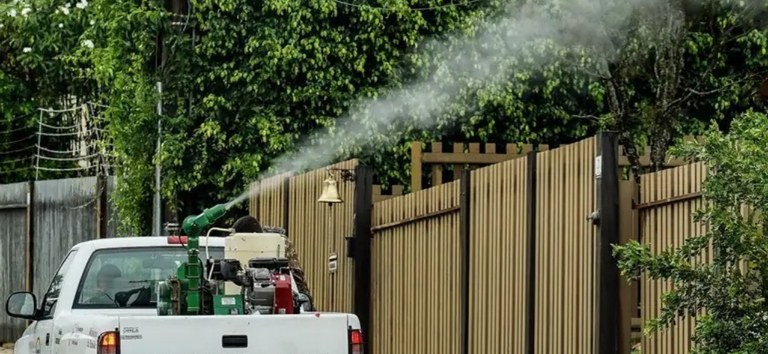 Carro fumacê lançando inseticida em névoa numa rua residencial