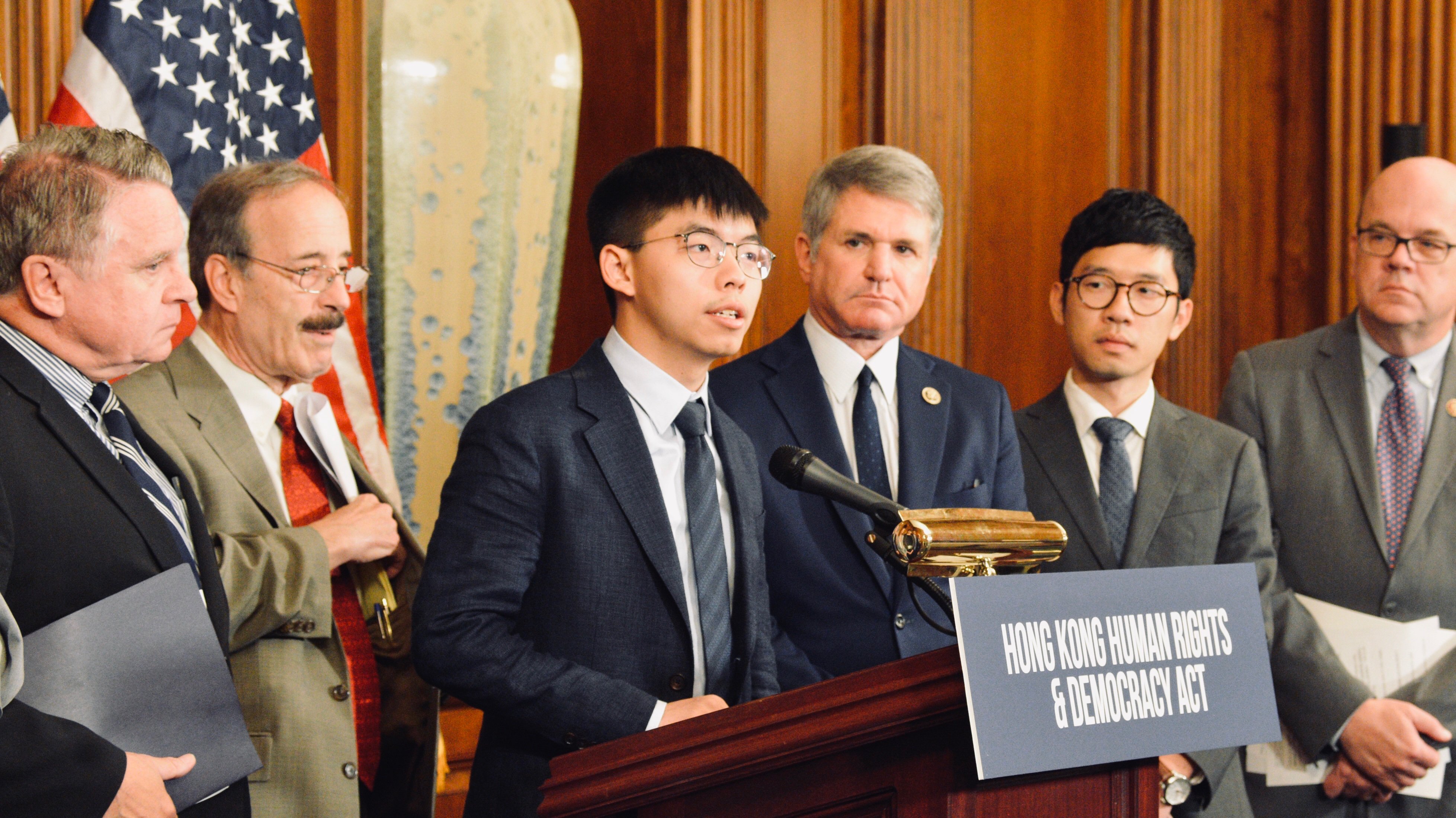 Joshua Wong discursando no Capitólio dos Estados Unidos em 2019, ao lado de Nathan Law e congressistas, com o letreiro Hong Kong Human Rights & Democracy Act.