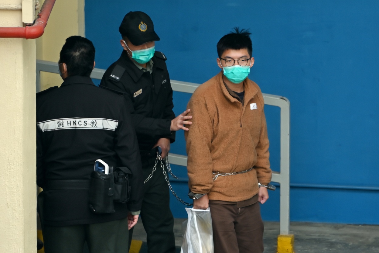 Joshua Wong escoltado pelo Departamento de Serviços Correcionais de Hong Kong, algemado, com máscara cirúrgica.
