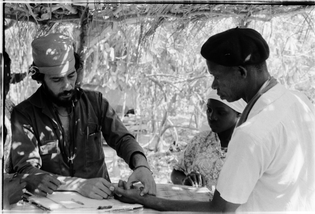 Médico cubano atendendo um paciente sentado, numa consulta improvisada durante missão internacional na Guiné-Bissau, em 1974.
