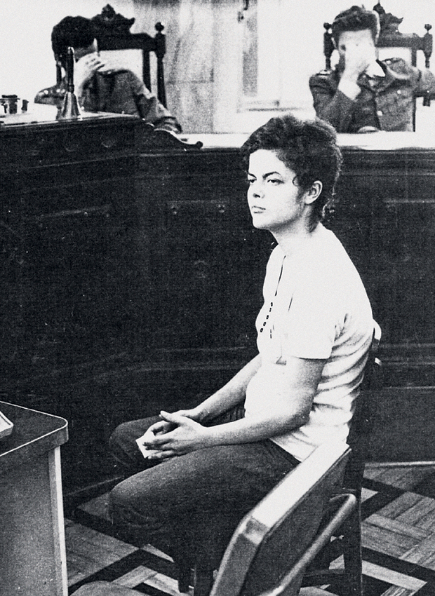 Dilma Rousseff sentada diante da Justiça Militar em 1970, enquanto dois oficiais ao fundo cobrem o rosto com as mãos.
